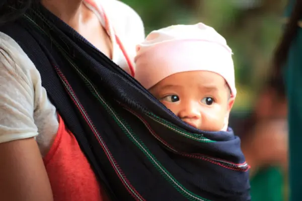 Bebe con gorrito recostado contra el pecho de su madre en una bandolera portabebe de tela sobre un hombro