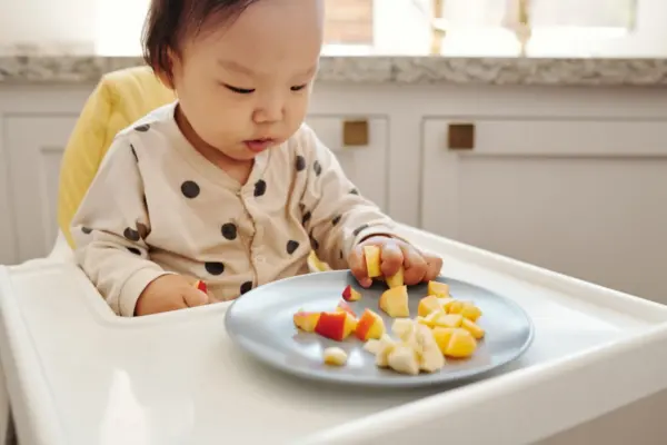 Bebé sentado en la trona comiendo trozos de fruta de un plato infantil apoyado en la bandeja