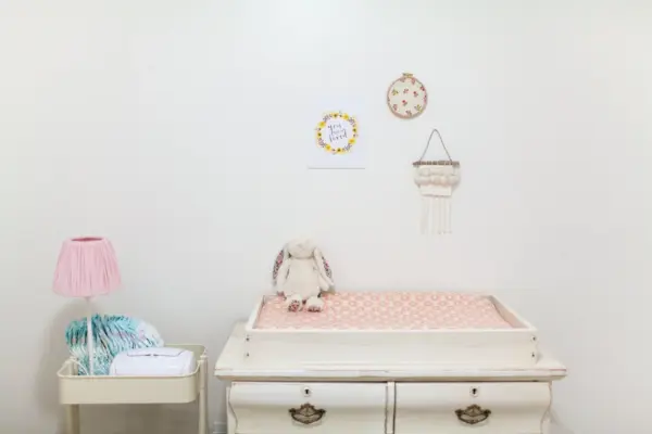Cambiador de bebé sobre una cómoda blanca en una habitación infantil luminosa, con un conejo de peluche encima y pañales en un carrito auxiliar